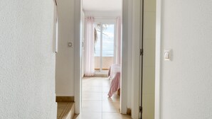 Corredor que dá para um quarto com cama, janela com cortinas e vista para a praia.