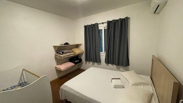 A bedroom with a bed, a shelf with folded clothes, a window with curtains, and a wall-mounted air conditioner.
