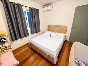 A bedroom with a bed, a window with curtains, an air conditioning unit, and a wooden floor. - Residencial Tristão de Oliveira (Gramado)