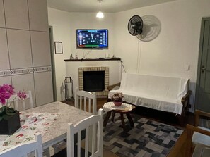 A living room with a television mounted on the wall, a fireplace, a dining table with chairs, and a small side table with a plant. - Residencial Tristão de Oliveira (Gramado)