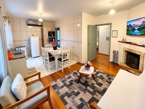 A modern living room with a fireplace, a dining area with a table and chairs, a kitchen with a refrigerator and stove, and a hallway leading to another room. - Residencial Tristão de Oliveira (Gramado)