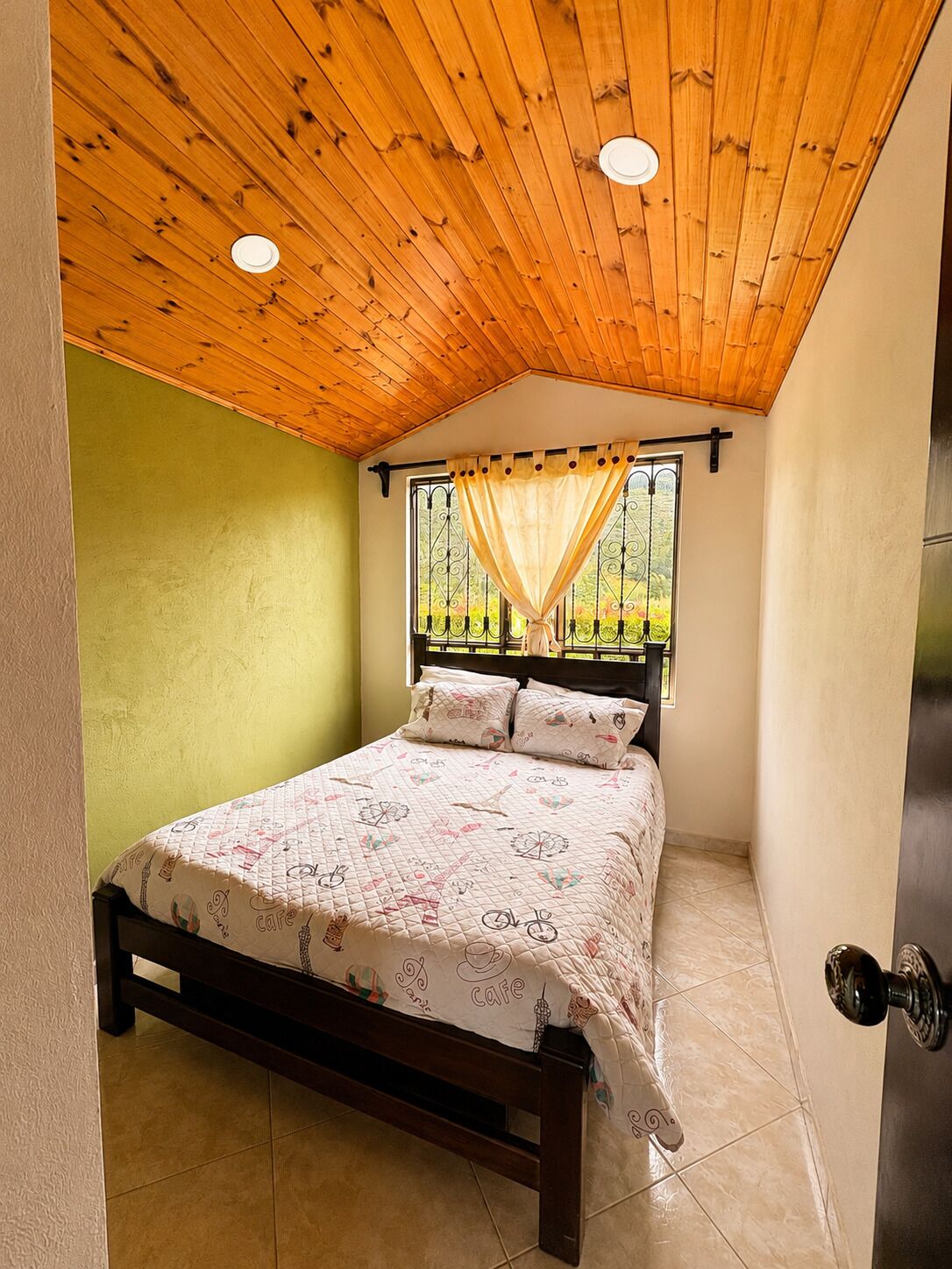 Un dormitorio con techo de madera, una cama con colcha estampada, una ventana con cortina y una pared verde.