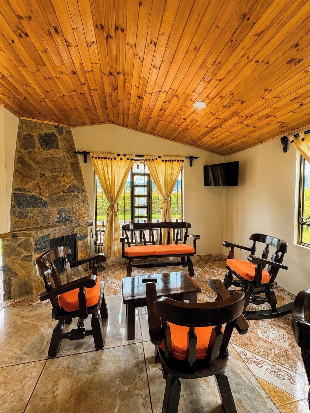 A room with a stone wall, wooden ceiling, and a stone fireplace. There are four wooden chairs with orange cushions and a wooden table. A television is mounted on the wall.