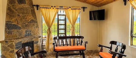 A room with a stone wall, wooden ceiling, and a stone fireplace. There are four wooden chairs with orange cushions and a wooden table. A television is mounted on the wall.