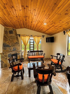 Um quarto com parede de pedra, teto de madeira e lareira de pedra. Há quatro cadeiras de madeira com almofadas laranja e uma mesa de madeira. Uma televisão está fixada na parede.