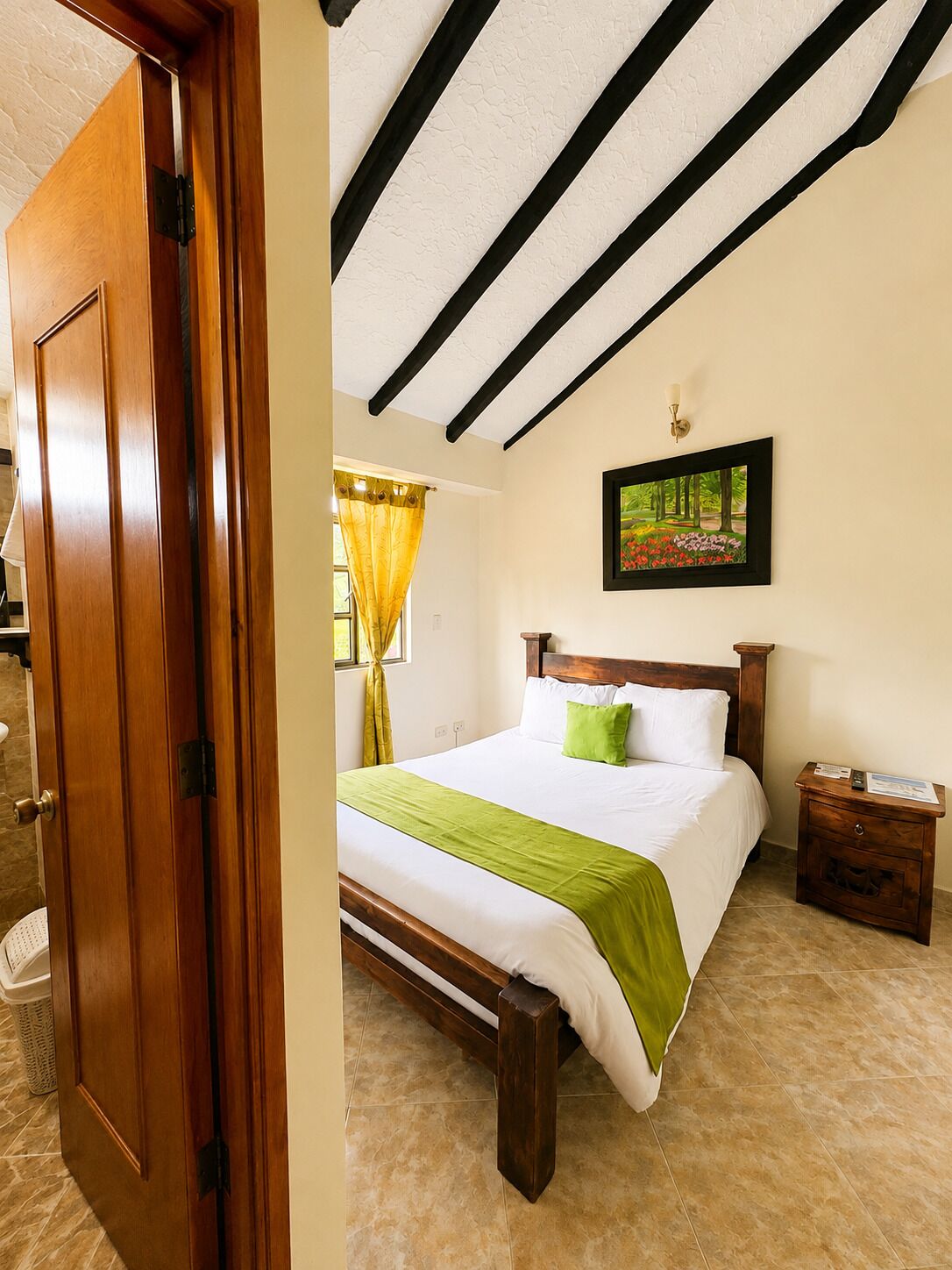 A hotel room with a wooden bed, a green throw, a painting on the wall, and a nightstand with a remote control.