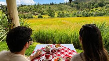 Um casal jantando à mesa com toalha quadriculada, em meio a uma paisagem verdejante e sob um céu claro.