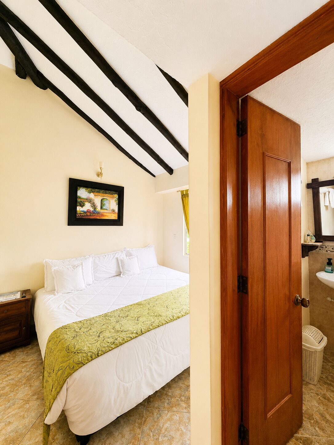 Une chambre d’hôtel avec un lit, une table de chevet, un tableau au mur, une salle de bain avec un lavabo et un miroir, et une porte donnant sur une autre pièce.