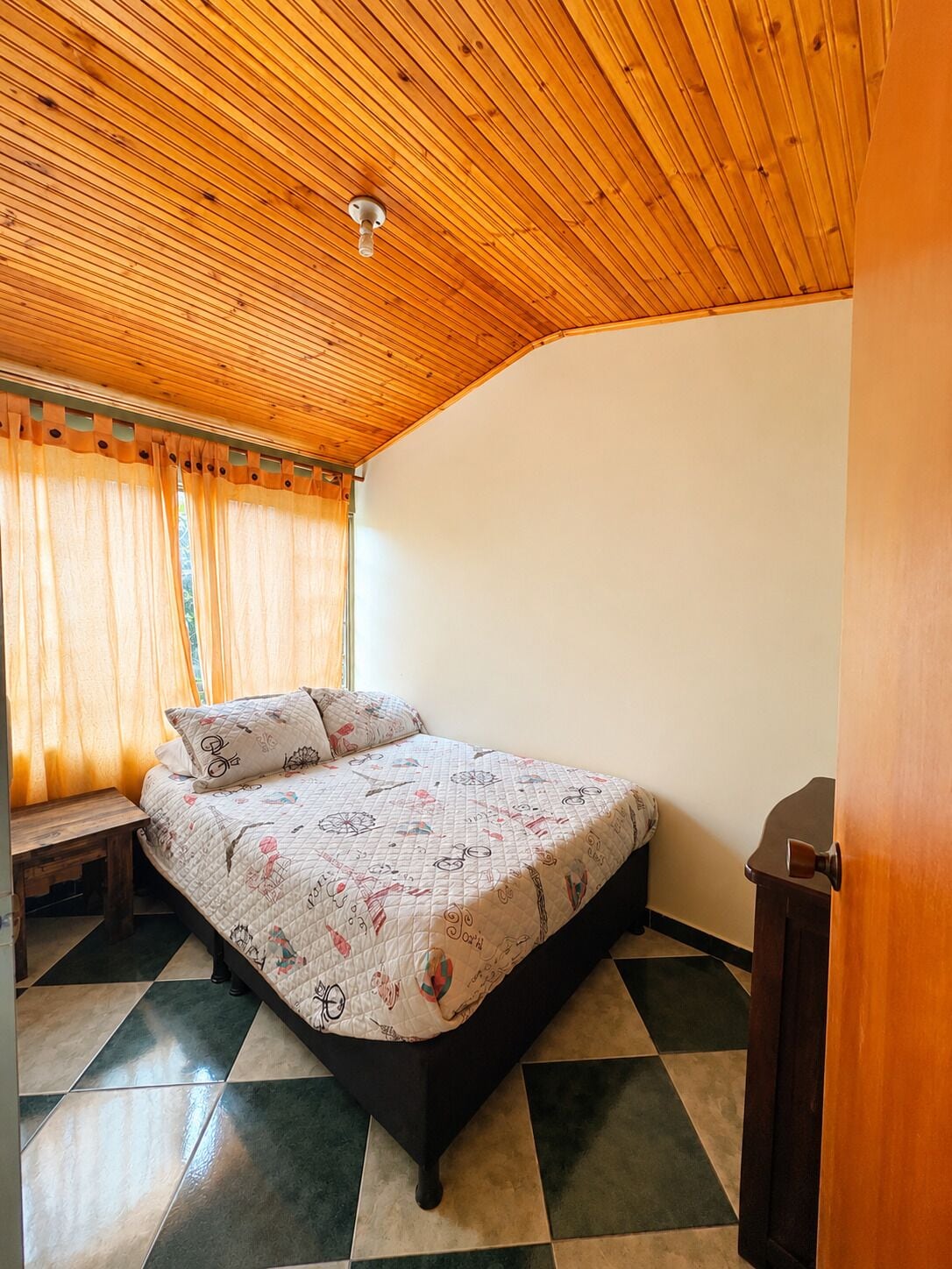 A bedroom with a wooden ceiling, a bed with a patterned quilt, a bench, and a door.