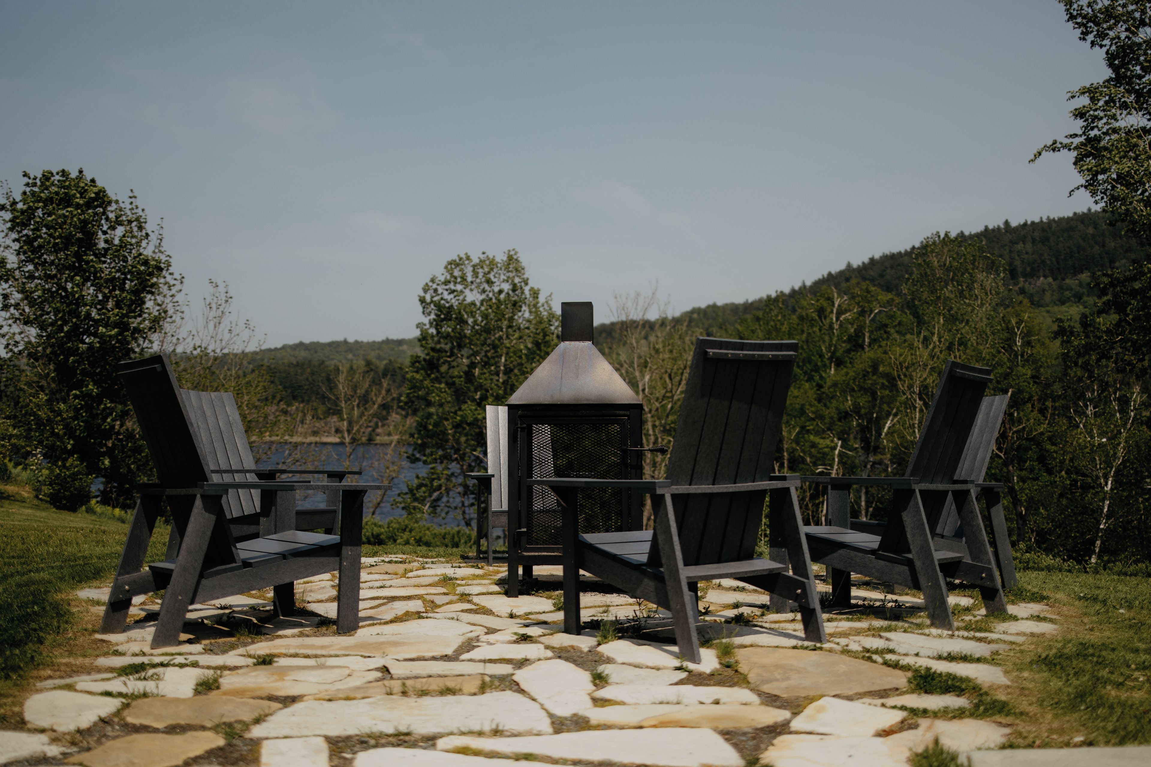 Tres sillas Adirondack dispuestas alrededor de un hogar de piedra en un entorno natural, con un lago y un bosque al fondo.