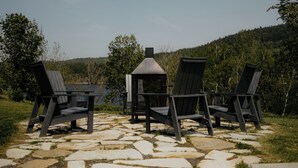 Tre Adirondack-stole placeret omkring et ildsted af sten i et naturområde med en sø og skov i baggrunden.