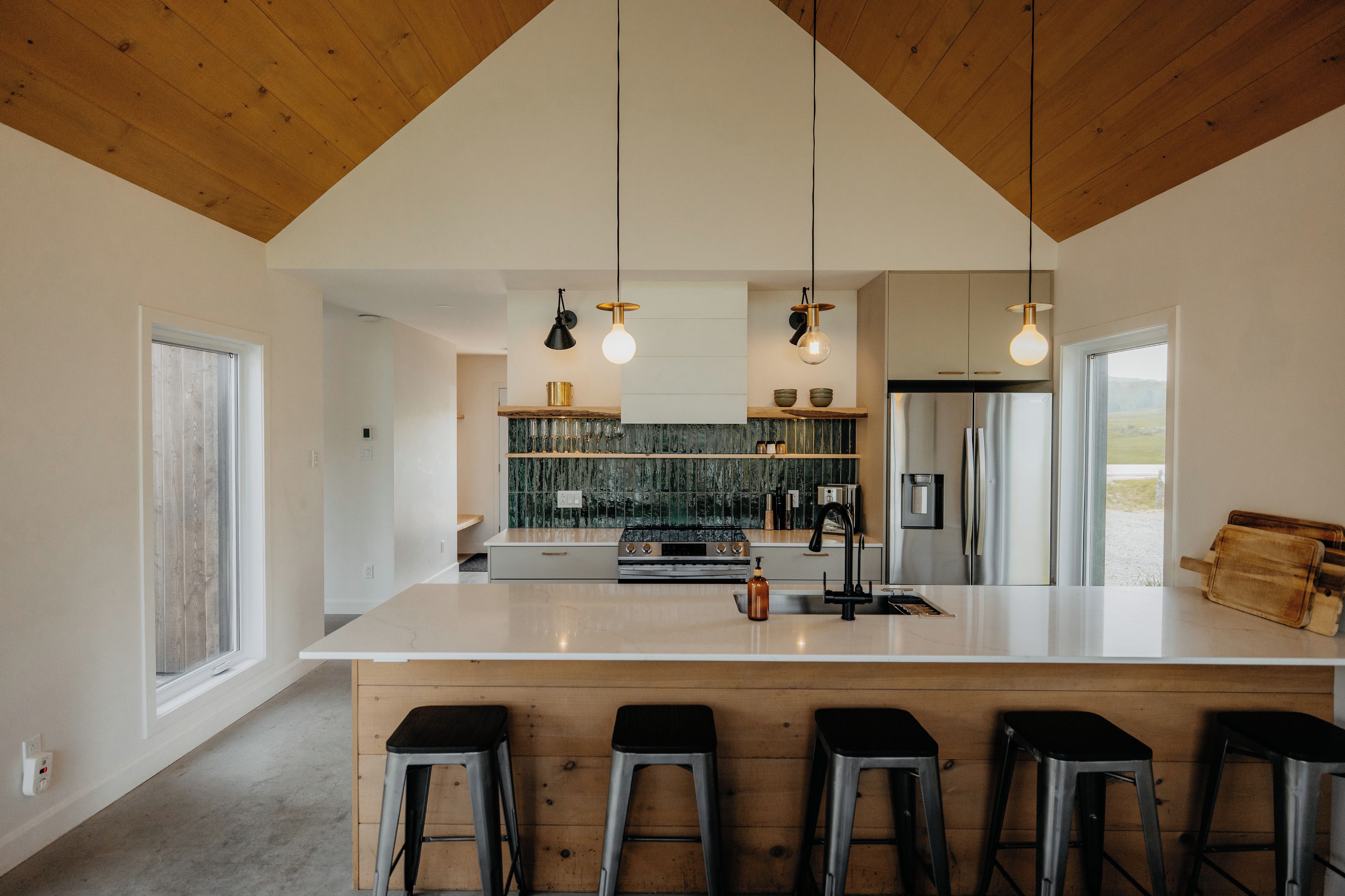 A modern kitchen with a large island, bar stools, hanging pendant lights, and a view of the outdoors.