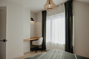 Un dormitorio con cama, escritorio, silla, lámpara y ventana con cortinas.