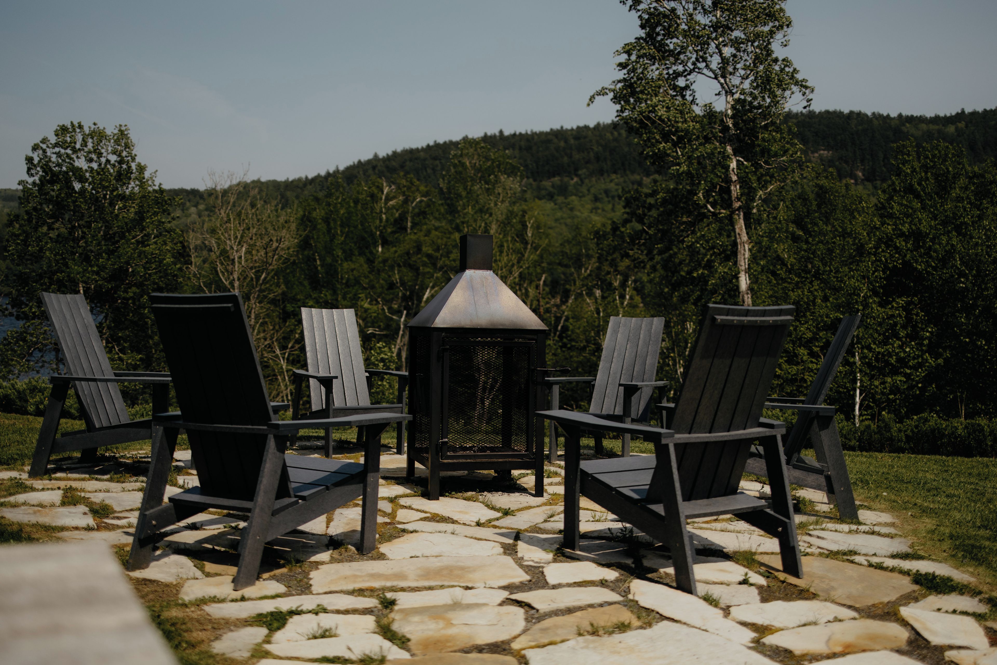 Outdoor seating with Adirondack chairs and a metal lantern, set on a stone patio with a forested hill in the background.