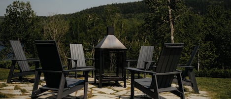 Outdoor seating with Adirondack chairs and a metal lantern, set on a stone patio with a forested hill in the background.
