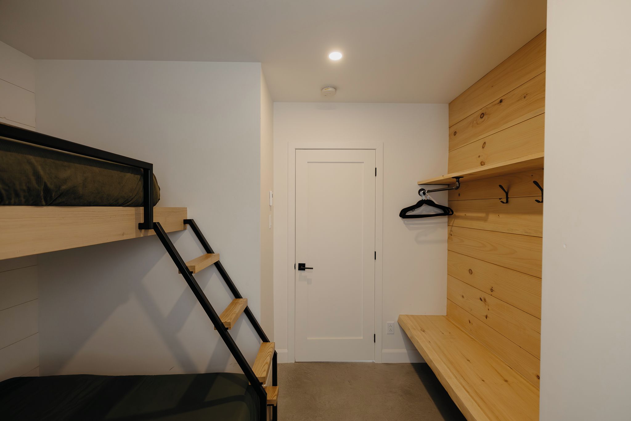 Una habitación con una cama litera, un banco de madera, una puerta y un estante con ganchos.