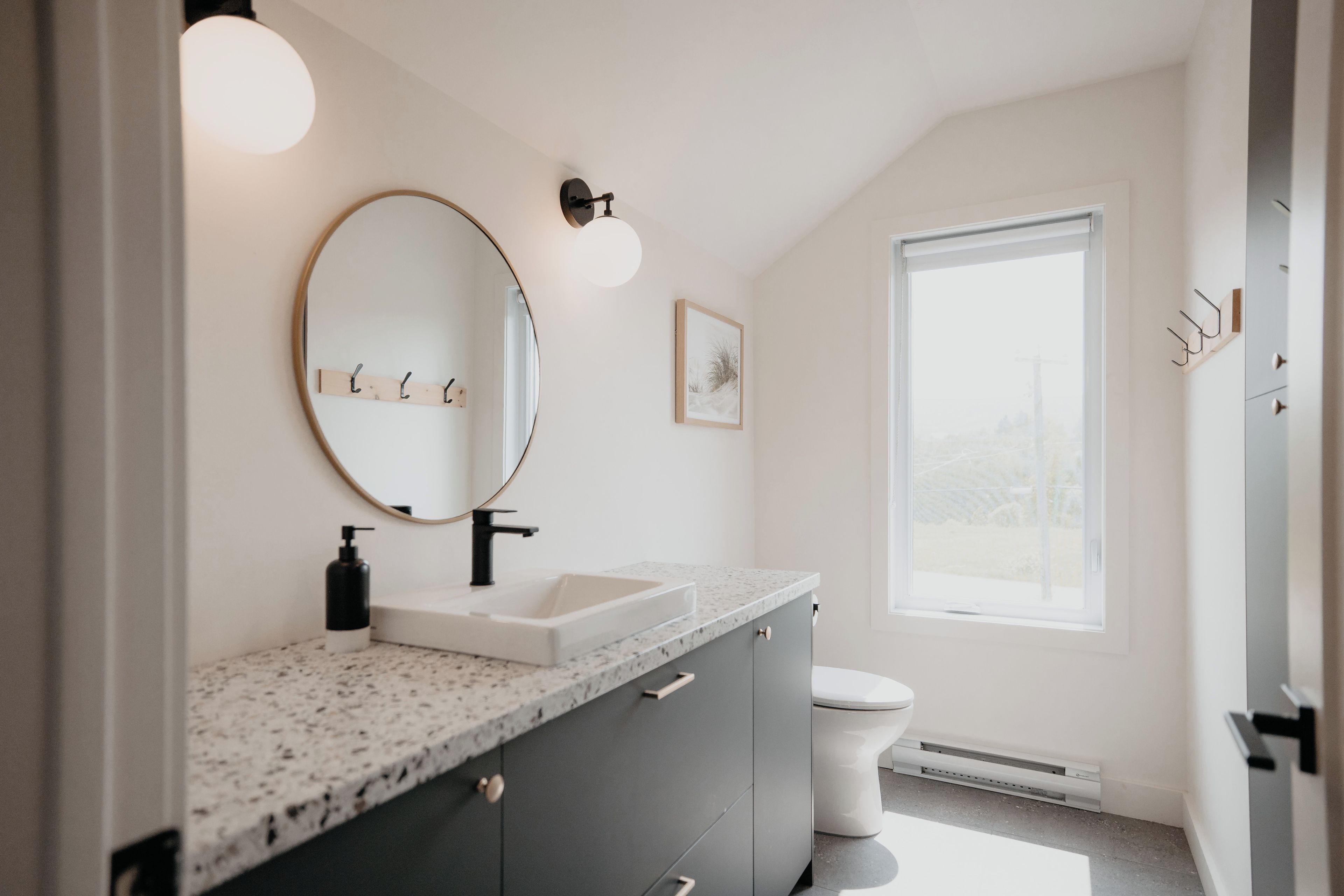 A modern bathroom with a granite countertop, a round mirror, a white sink, a toilet, and a window providing natural light.