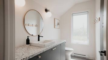 A modern bathroom with a granite countertop, a round mirror, a white sink, a toilet, and a window providing natural light.