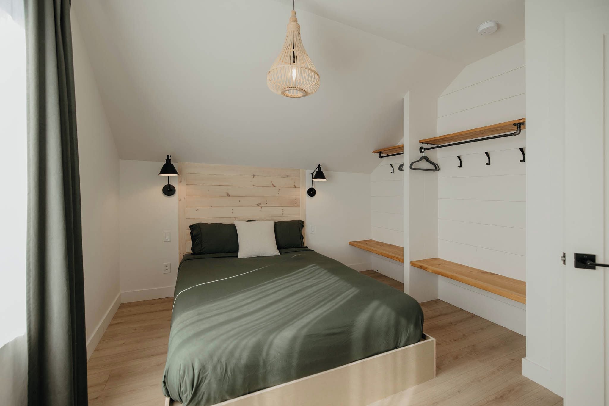 A bedroom with a bed, wooden headboard, bedside lamps, a hanging light fixture, a wardrobe, and a wooden shelf.