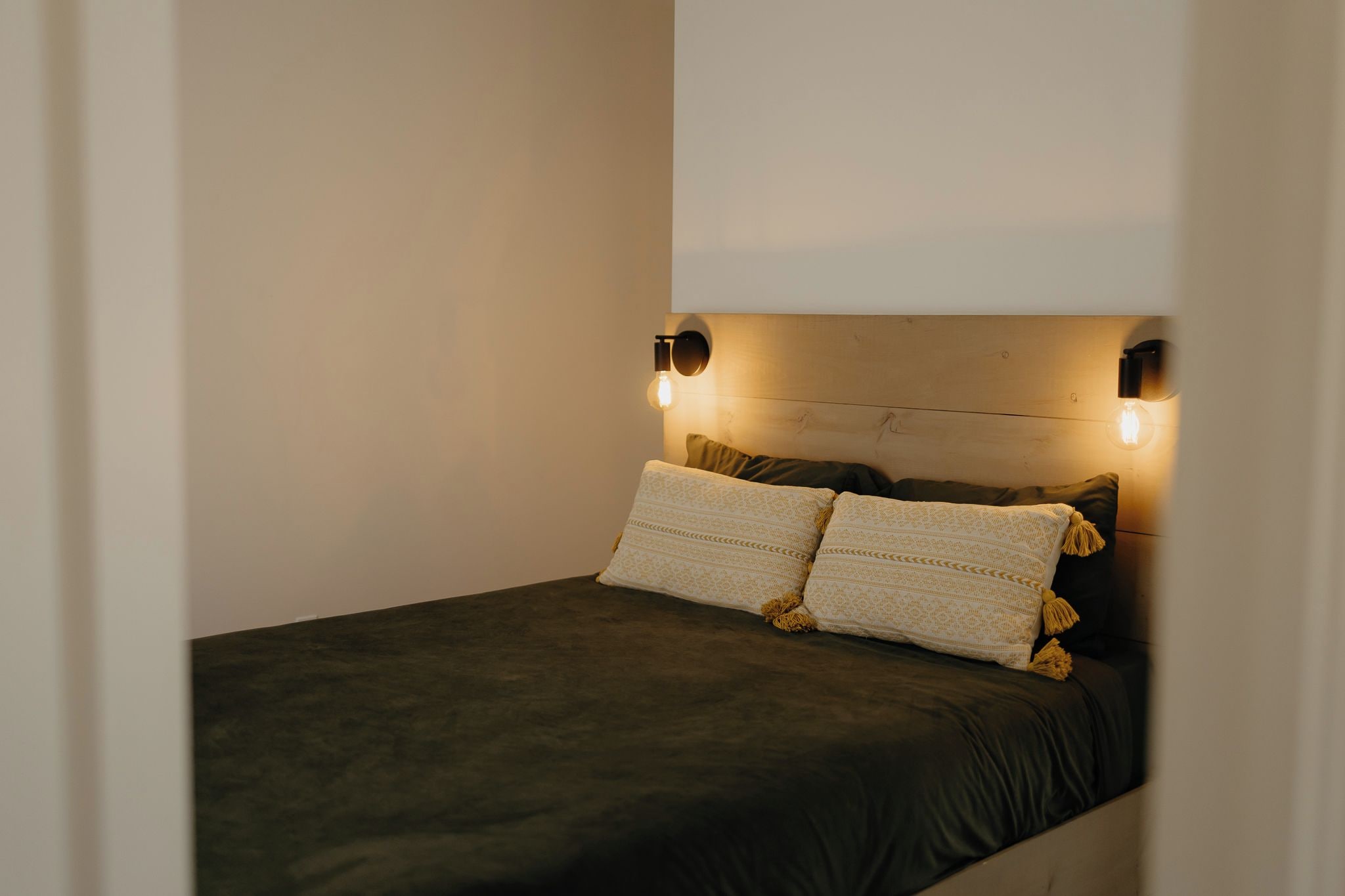 Una cama individual con colchón oscuro, dos almohadas y cabecera con luces fijadas a la pared.