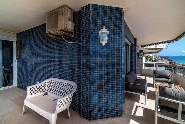 Un balcon avec un mur recouvert de carreaux bleus, une chaise blanche, une table en bois et un luminaire blanc suspendu.