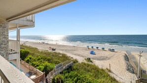 Blick auf den Strand vom Balkon aus, mit Pool, Strandliegen und blauem Himmel.