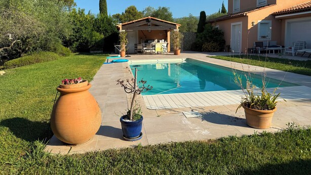 A swimming pool with a patio area, a small covered seating area, and a house with a tiled roof.