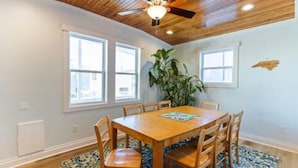 Une salle à manger avec une table et des chaises en bois, un ventilateur de plafond et de grandes fenêtres.