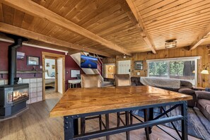 A rustic cabin interior with a wooden dining table, a fireplace, a sofa, and a large window. - Sawmill Trail Tahoe Cabin (South Lake Tahoe)