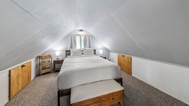 Ein Schlafzimmer mit schrägen Wänden, einem Bett mit weißer Bettwäsche, einem Nachttisch mit einer Lampe, einer Holzbank und einem kleinen Fenster mit Vorhängen.