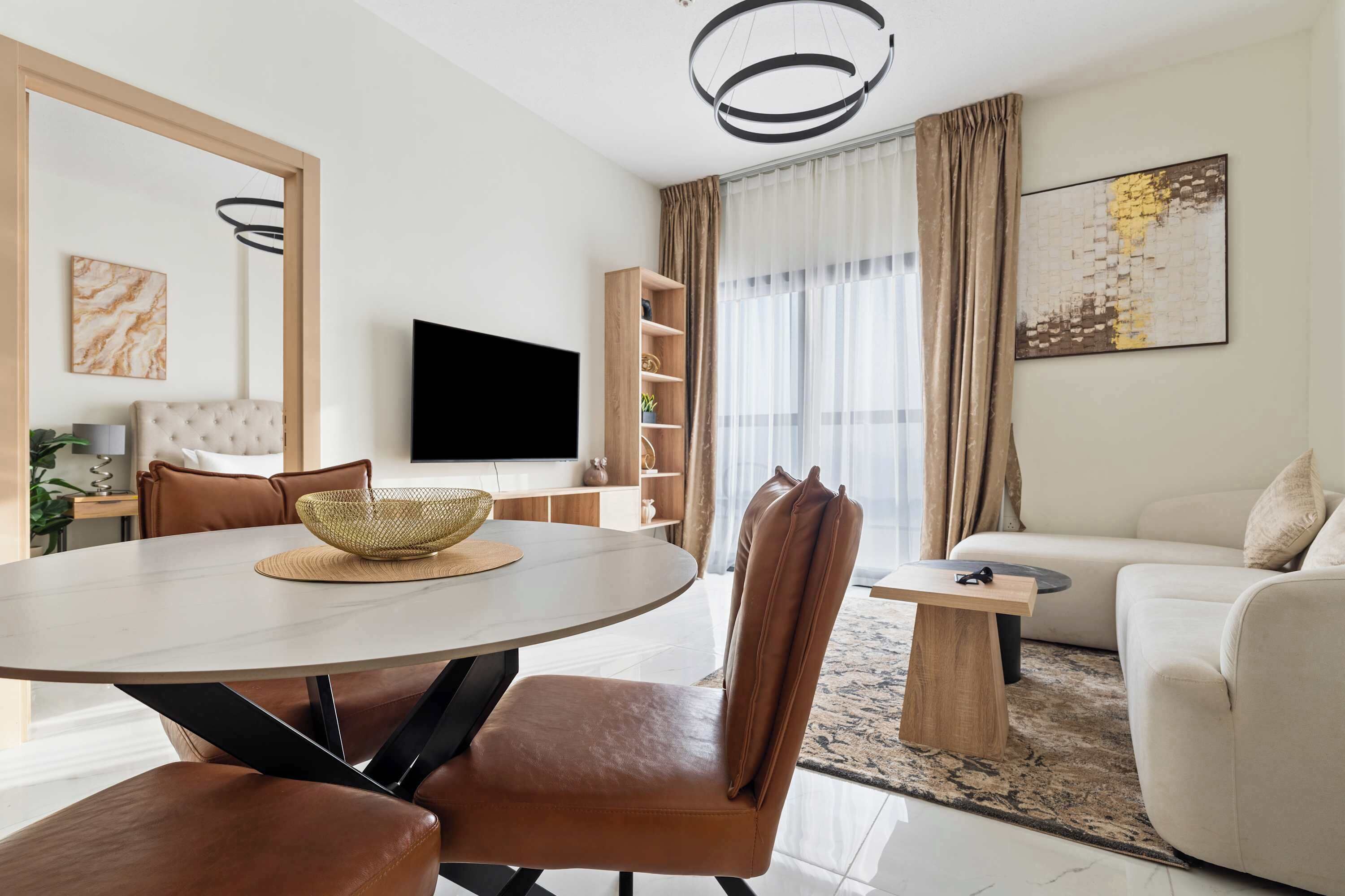 A modern living room with a round dining table, leather chairs, a sofa, a flat-screen TV, and a bookshelf.