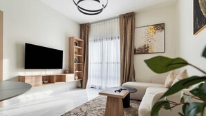 Ein modernes Wohnzimmer mit Flachbildfernseher, Couch, Holz-Couchtisch, Bücherregal und einem großen Fenster mit Vorhängen.