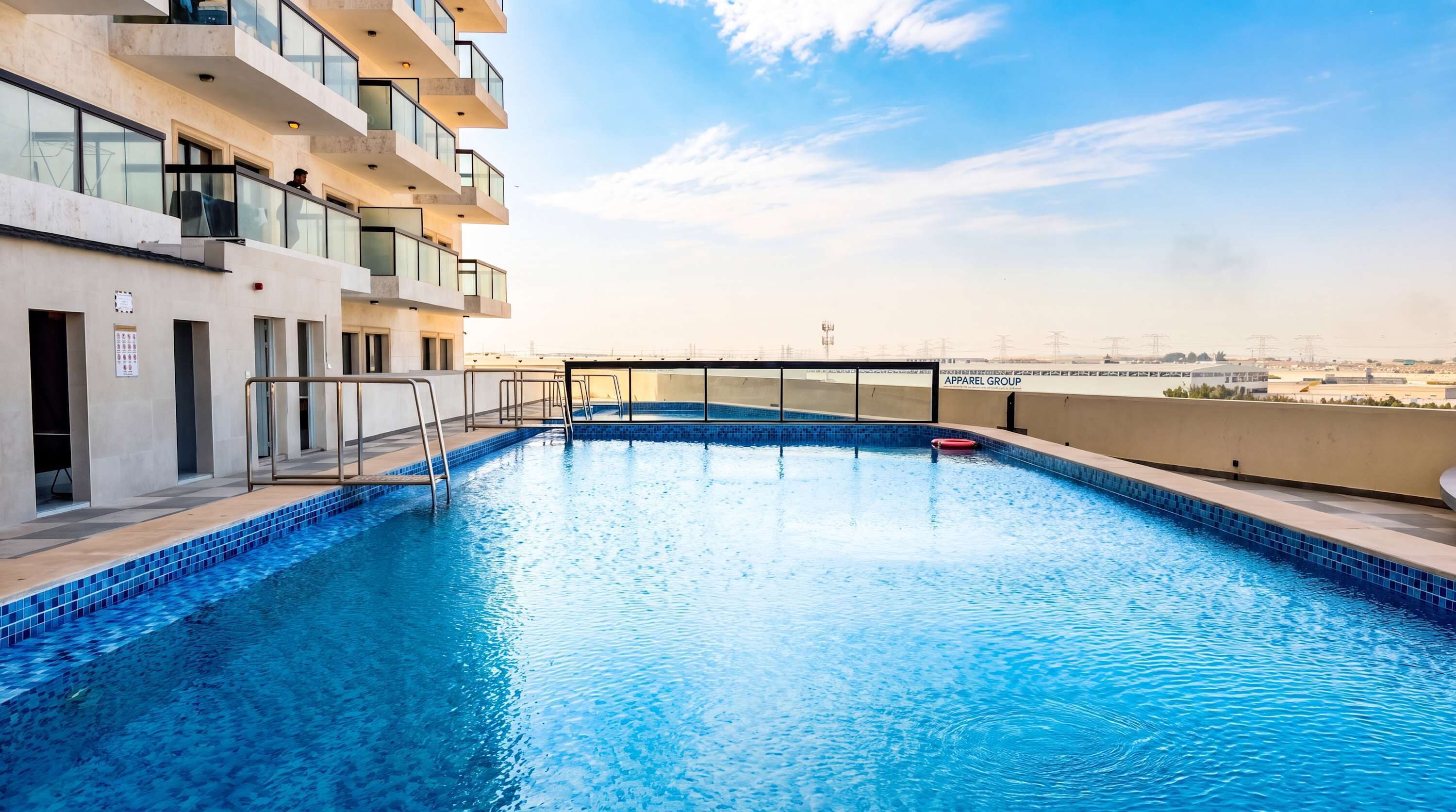 A rooftop swimming pool with a clear view of the city skyline, a modern building with balconies, and a sign for 'APPAREL GROUP'.