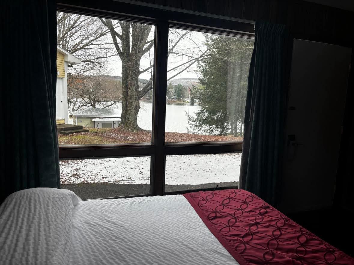 Una habitación con una cama, una ventana que da a un lago y casas, y una puerta.