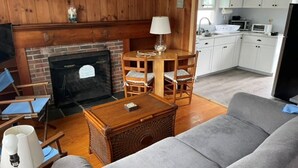 Una sala de estar con chimenea, un sofá, una mesa de comedor con sillas y una zona de cocina con armarios blancos.