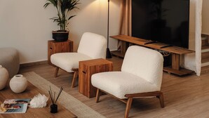 Ein modernes Wohnzimmer mit zwei weißen, flauschigen Sesseln, einem Holztisch, einer Stehlampe, einem großen Flachbildfernseher, einer Topfpflanze und einer Zeitschrift auf dem Boden.