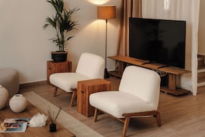 Un salon moderne avec deux fauteuils blancs et moelleux, une table basse en bois, un lampadaire, un grand téléviseur à écran plat, une plante en pot et un magazine posé sur le sol.