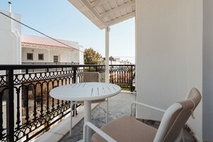 Un balcón con una mesa y sillas blancas, baranda de hierro forjado negro, y vista de edificios al fondo.