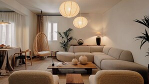 Ein modernes Wohnzimmer mit einer Couch, zwei Sesseln, einem Couchtisch und einem hängenden Rattanstuhl. Auf einem Beistelltisch steht eine Lampe und es gibt einen Topfpflanzen.