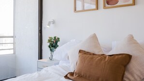 Ein Schlafzimmer mit einem Bett, Kissen, einem Nachttisch, einer Vase mit Blumen und zwei an der Wand befestigten abstrakten Gemälden.