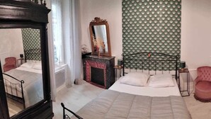 A bedroom with a bed, a mirror, a fireplace, and a patterned wall. - The Cozy Baroque Nest—Linens Provided (La Bourboule)