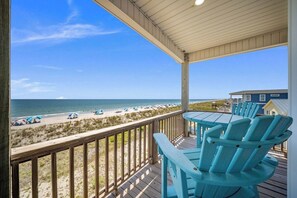 A wooden deck with a view of the beach, a table with chairs, and a building in the background. - Paradis Marin | Oceanfront + Private Pool (Oak Island)
