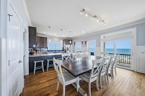 A spacious dining area with a large table and chairs, a kitchen with a bar, and a view of the ocean through large windows. - Paradis Marin | Oceanfront + Private Pool (Oak Island)
