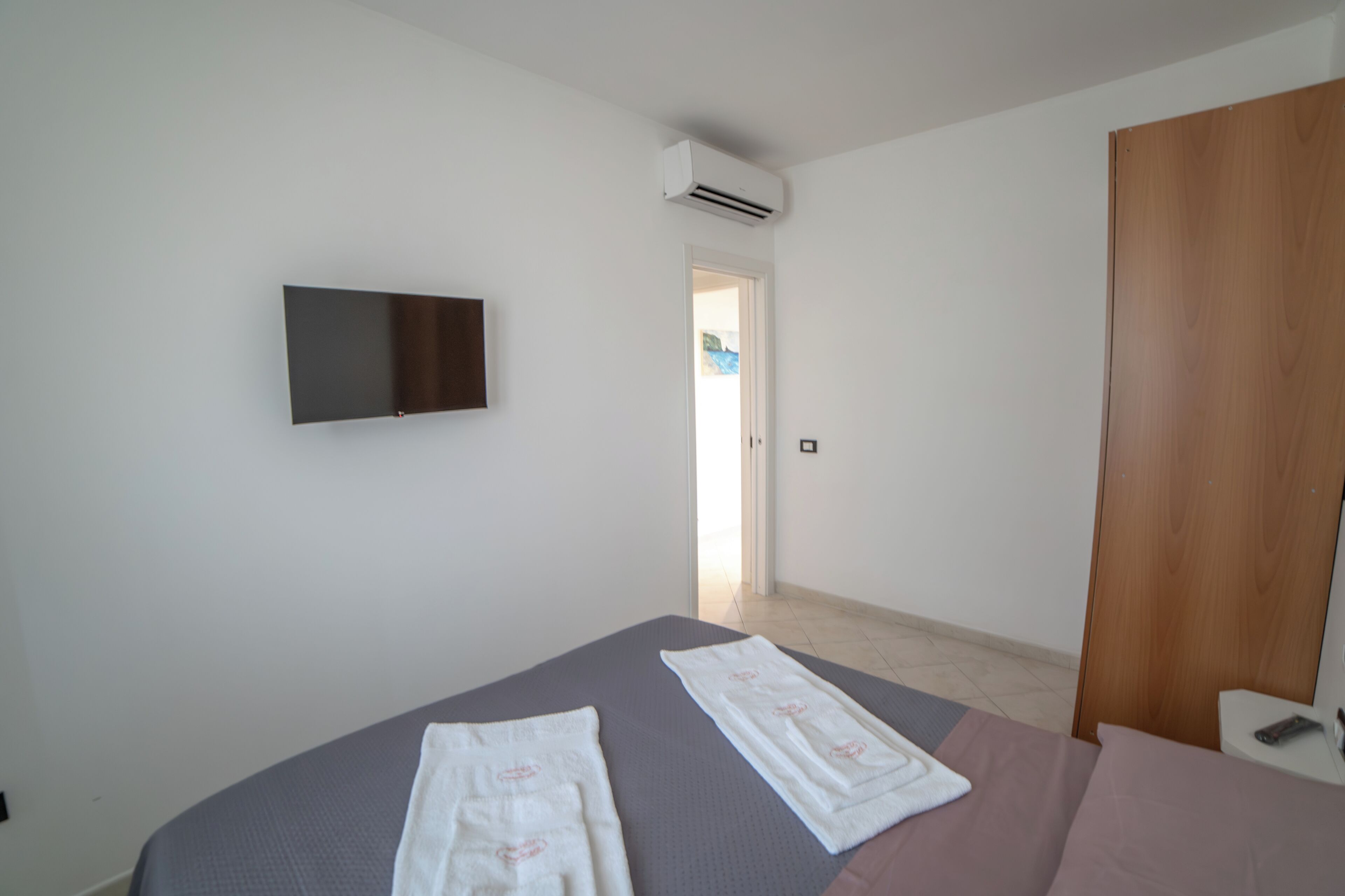 A bedroom with a bed, a flat-screen TV mounted on the wall, an air conditioning unit, and a wooden door.