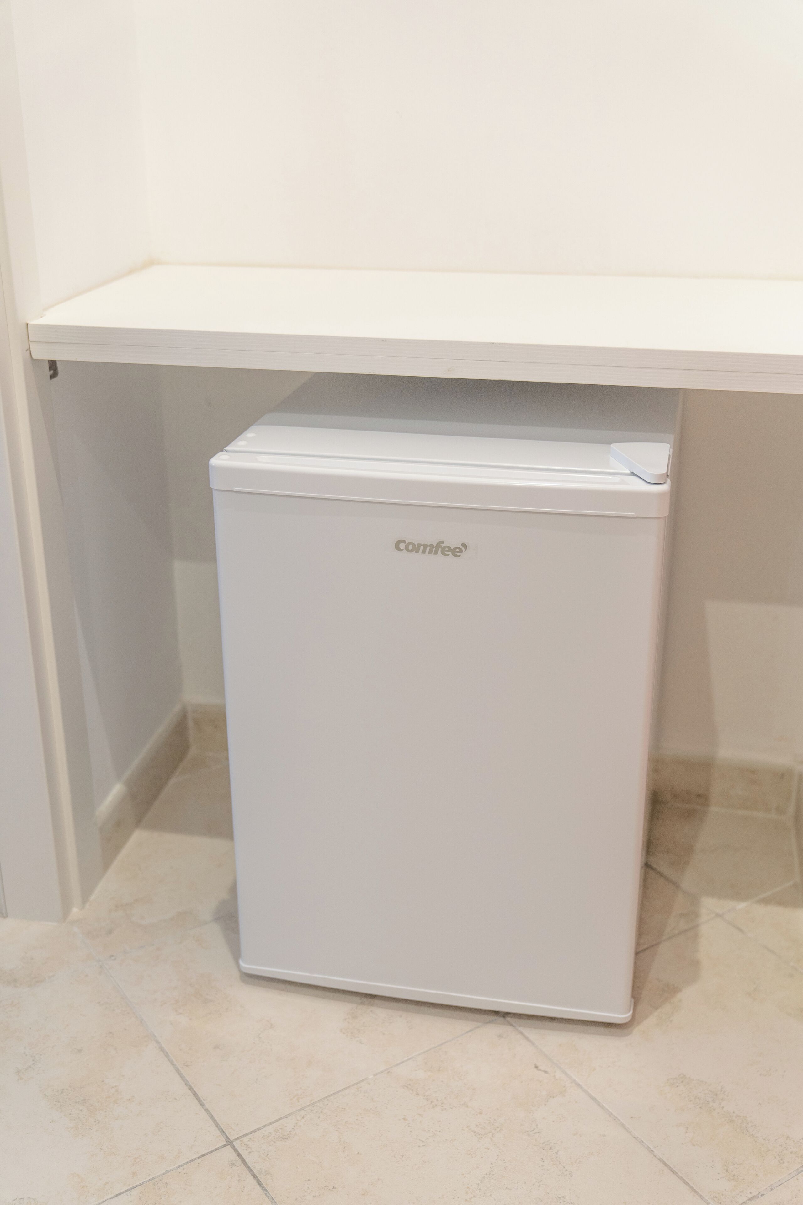 Un refrigerador Comfee blanco compacto en un rincón con un estante blanco encima.