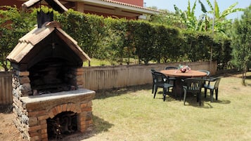 Un horno de ladrillo al aire libre con chimenea, rodeado por una mesa y sillas de madera, con una casa y abundante vegetación de fondo.