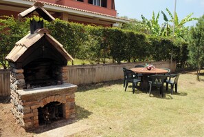 An outdoor brick oven with a chimney, surrounded by a wooden table and chairs, set against a backdrop of a house and lush greenery.
