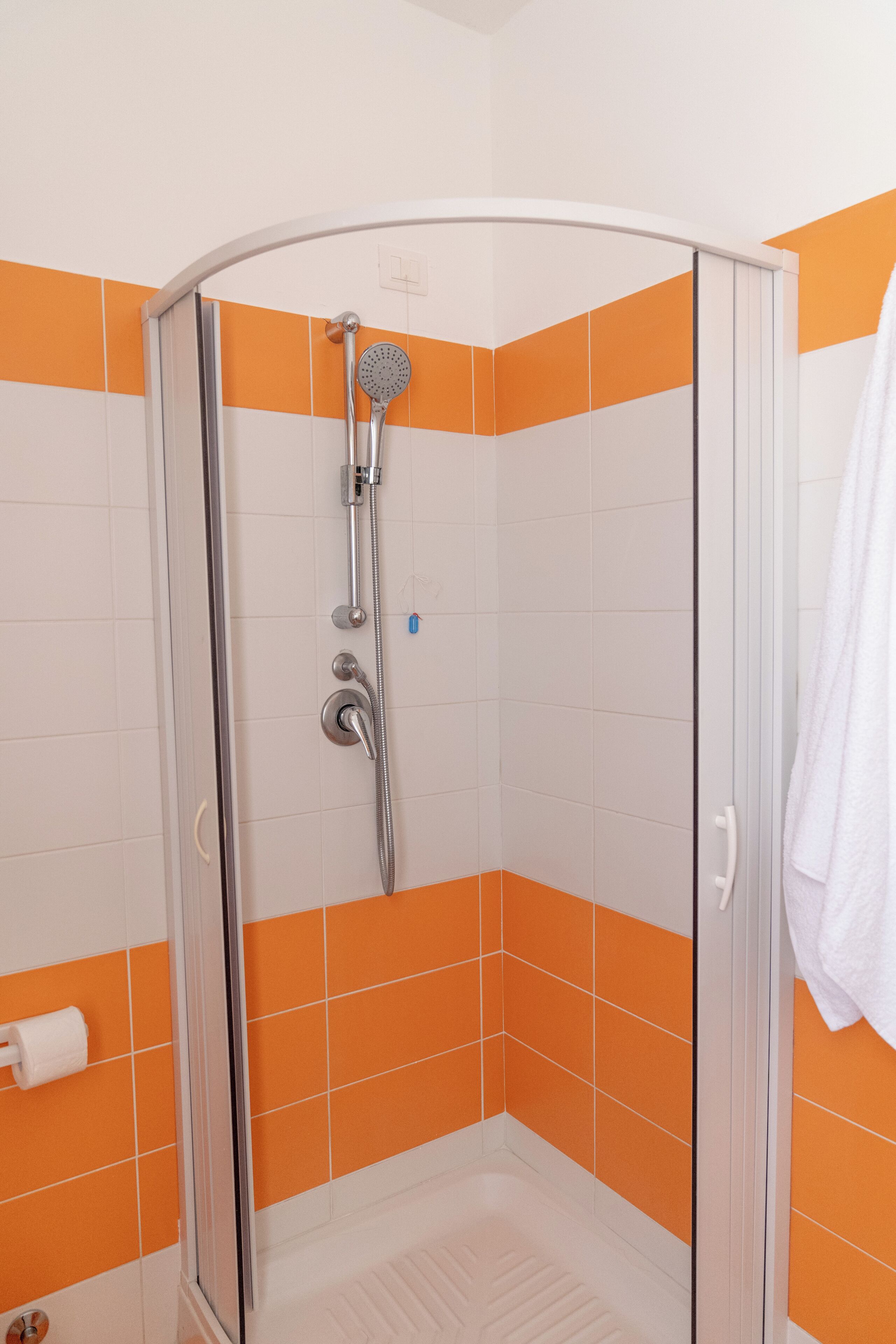 Chuveiro com porta de vidro, acessórios cromados e paredes azulejadas em tons de laranja e branco.