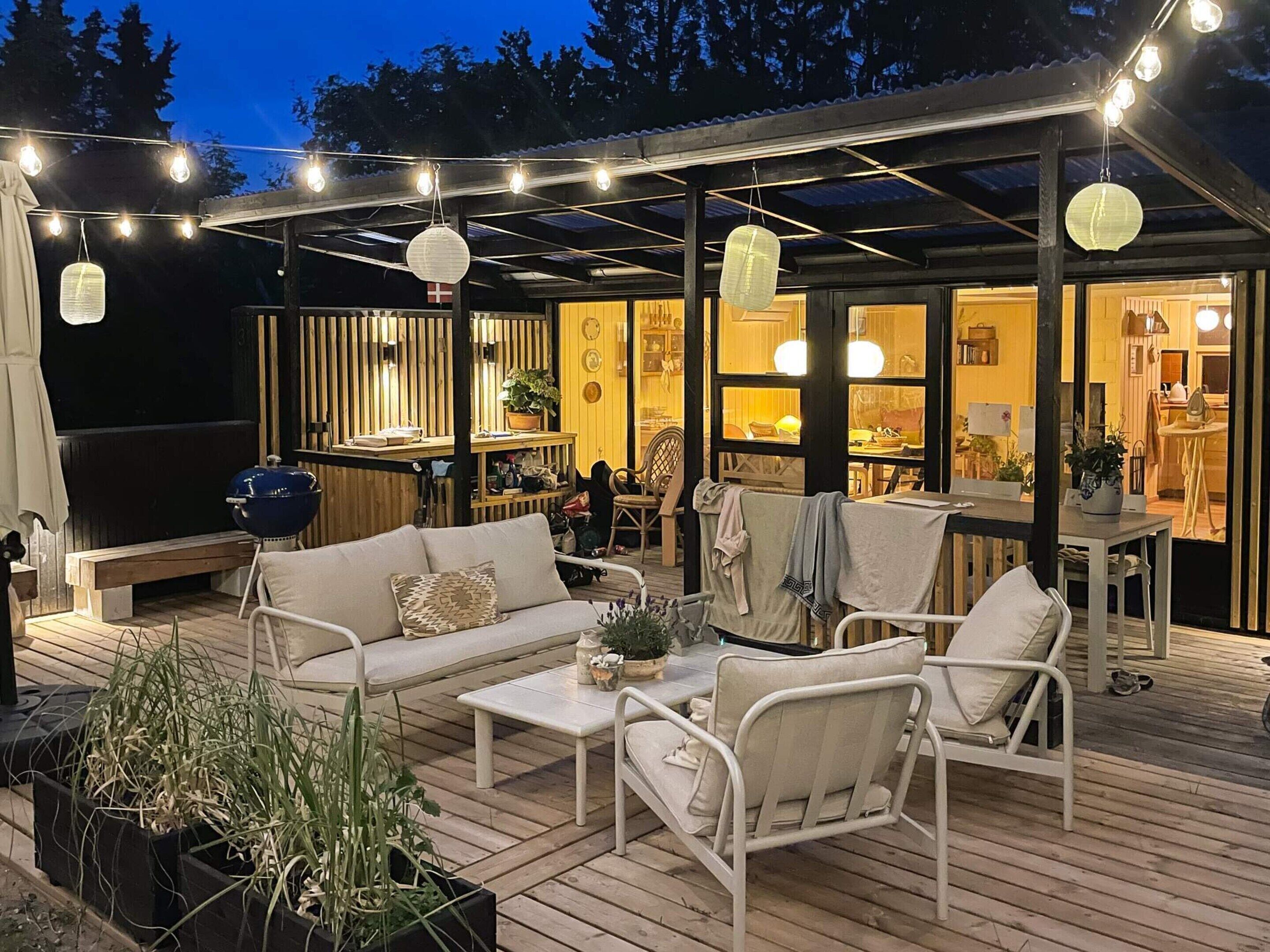 An outdoor deck with a sofa, chairs, and a table, connected to a building with a kitchen visible through large windows.
