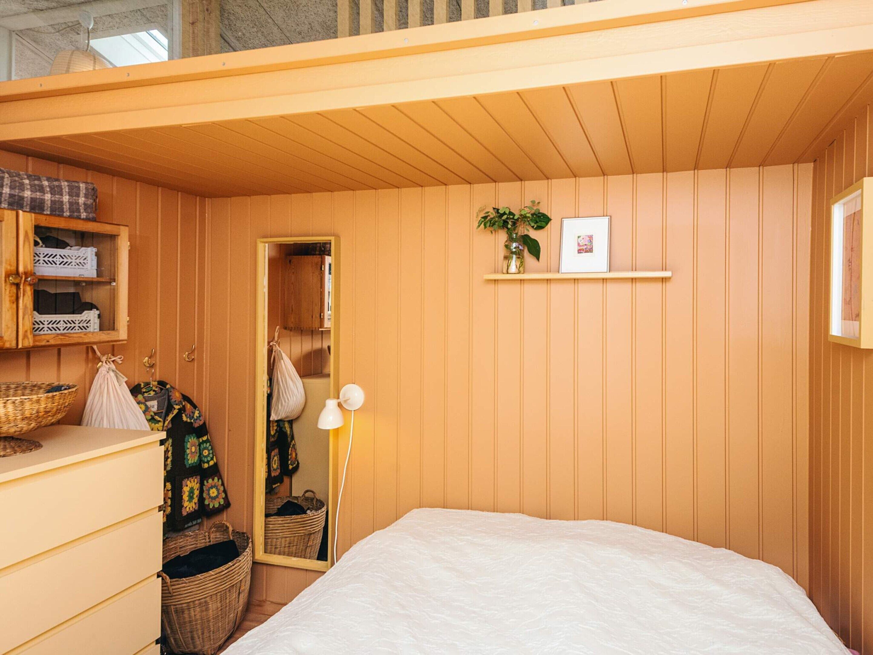 A bedroom with a bed, dresser, mirror, and a shelf with a plant and picture.